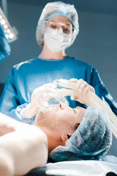 Селективный фокус медсестры в форме и медицинской шапке, надевающей маску на пациента — стоковое фото