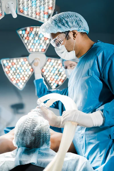 Selective focus of doctor in uniform and medical cap putting mask on patient putting — Stock Photo