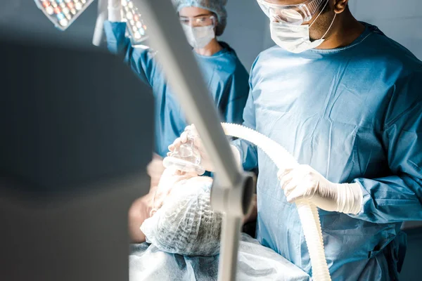 Selective focus of surgeon in uniform and goggles putting mask on patient — Stock Photo