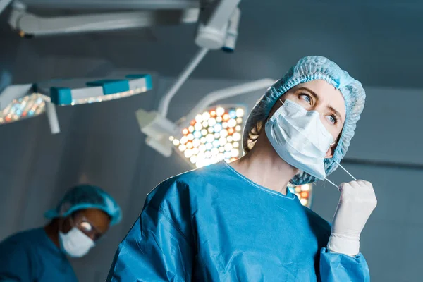 Selective focus of nurse in uniform putting off medical mask in operating room — Stock Photo