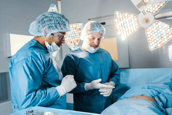 Enfermeiro e cirurgião em uniformes e máscaras médicas conversando na sala de cirurgia — Fotografia de Stock