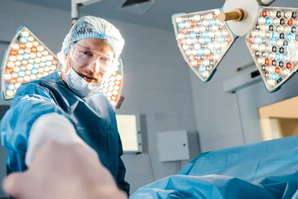 Selektiver Fokus des Chirurgen in Uniform und medizinischer Maske, der in die Kamera schaut und die Hand des Patienten hält — Stockfoto