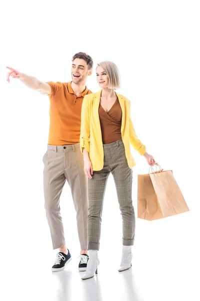 Hombre guapo señalando con el dedo, mientras que de pie cerca de la mujer bonita con bolsas de papel aisladas en blanco - foto de stock