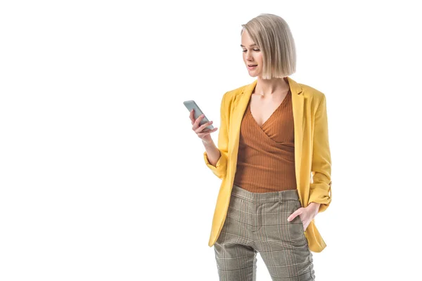 Atractiva mujer con la mano en el bolsillo sosteniendo teléfono inteligente aislado en blanco - foto de stock