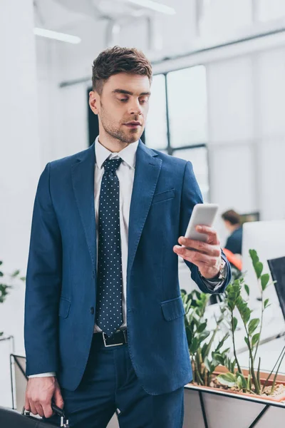 Hombre de negocios guapo y serio usando el teléfono inteligente en la oficina - foto de stock