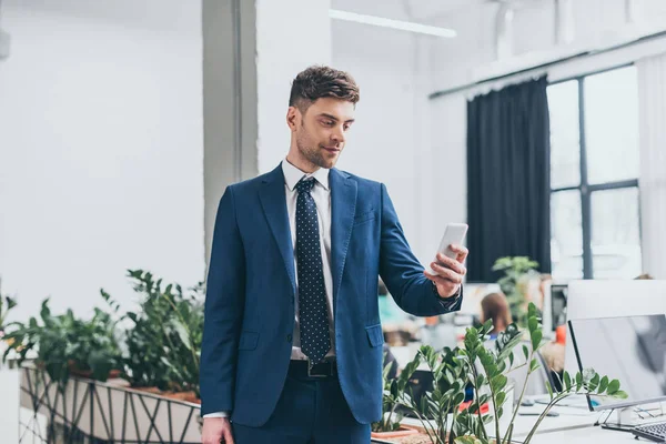 Hombre de negocios guapo y sonriente en suite con teléfono inteligente en la oficina — Stock Photo