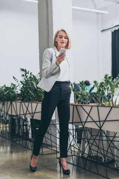 Pretty businesswoman in formal wear and with hand in pocket using smartphone in office — Stock Photo