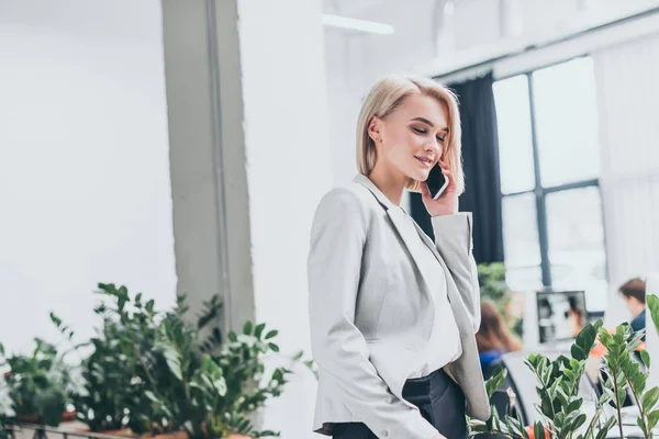 Bastante sonriente mujer de negocios en ropa formal hablando en el teléfono inteligente en la oficina - foto de stock