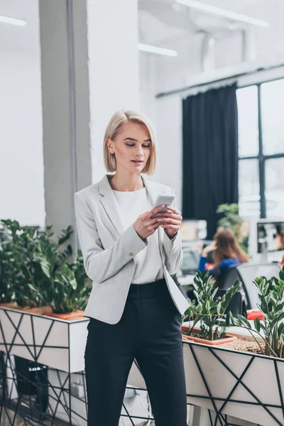 Красивая улыбающаяся деловая женщина в формальной одежде с помощью смартфона в офисе — стоковое фото