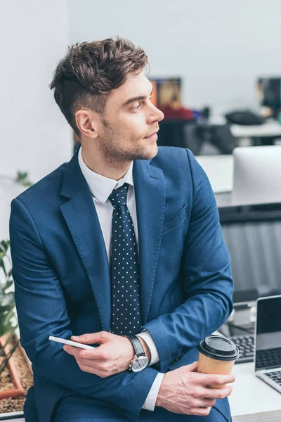 Bel homme d'affaires sérieux tenant smartphone et tasse en papier et détournant les yeux — Photo de stock