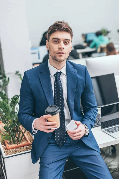 Homme d'affaires confiant tenant smartphone et tasse en papier et regardant la caméra — Photo de stock