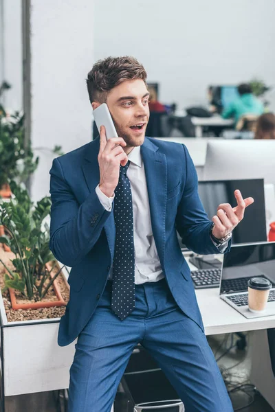 Cheerful businessman holding talking on smartphone and gesturing in office — Stock Photo