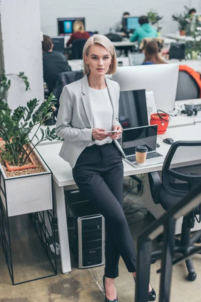 Pretty blonde businesswoman in formal wear standing by workplace and looking at camera — Stock Photo