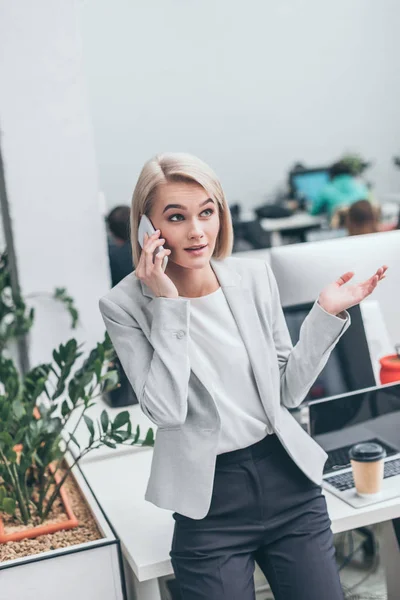 Attraktive lächelnde Geschäftsfrau, die im Büro mit dem Smartphone spricht und gestikuliert — Stockfoto