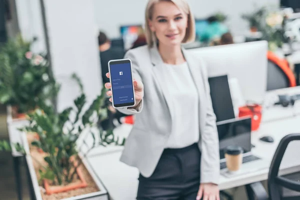 Selective focus of pretty businesswoman holding smartphone with facebook app on screen — Stock Photo