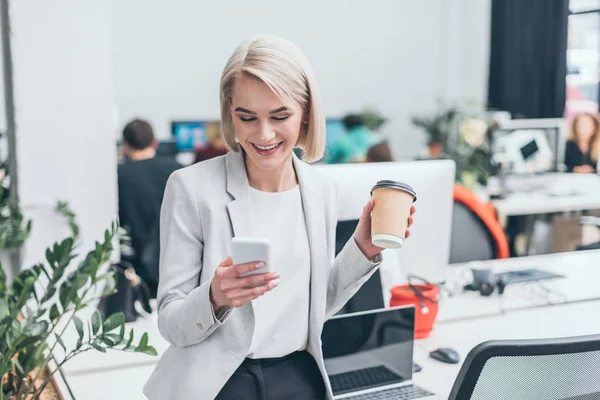 Bella donna d'affari sorridente utilizzando smartphone e tenendo tazza di carta in ufficio — Foto stock