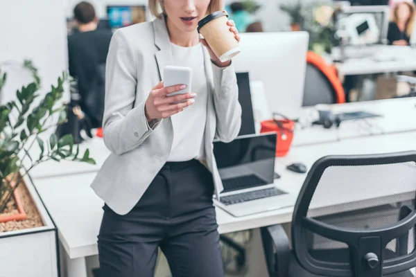 Partial view of businesswoman using smartphone and drinking coffee in office — Stock Photo