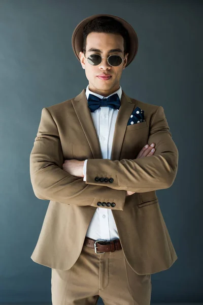 Élégant mixte homme en costume et lunettes de soleil posant avec les bras croisés isolés sur gris — Photo de stock