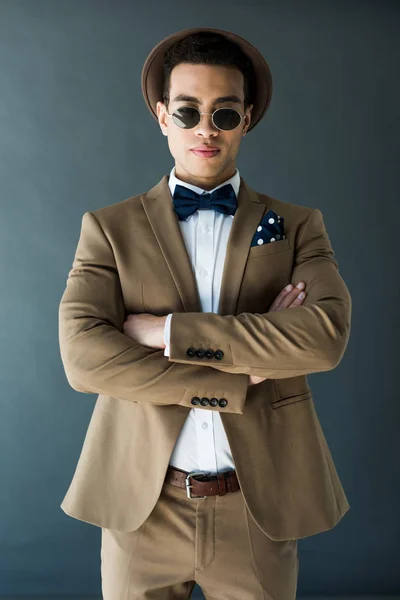 Elegante hombre de raza mixta en traje y gafas de sol posando con brazos cruzados aislados en gris - foto de stock