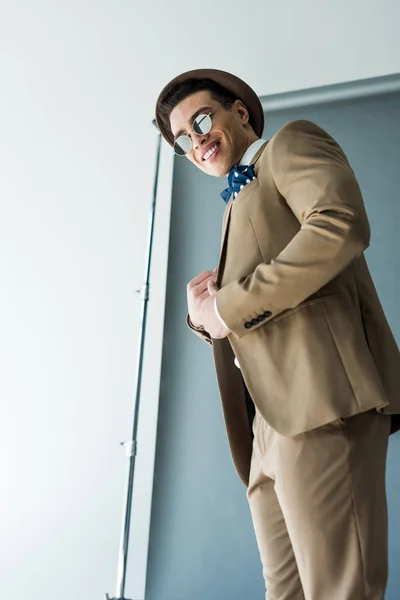 Fashionable mixed race man in suit and hat smiling and posing on grey with copy space — Stock Photo