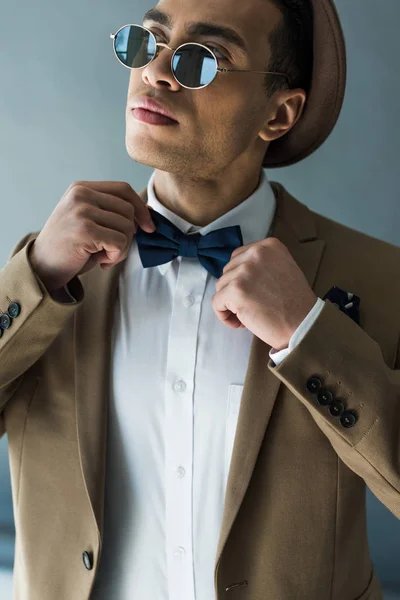 Elegante hombre de raza mixta en traje y gafas de sol ajustando pajarita en gris - foto de stock