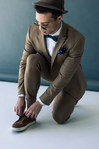 Elegante hombre de raza mixta en traje y gafas de sol que se pone el zapato en gris y blanco - foto de stock