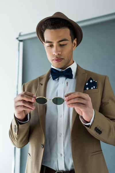 Stylish mixed race man in suit and bow tie holding sunglasses on grey — Stock Photo