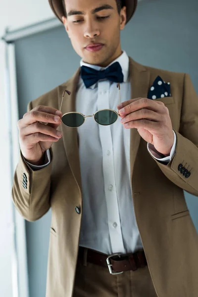 Élégant mixte homme en costume et noeud papillon tenant des lunettes de soleil sur gris — Photo de stock