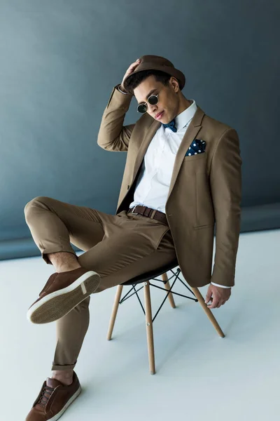 Elegante hombre de raza mixta en traje y gafas de sol sentado en la silla y posando en gris y blanco - foto de stock