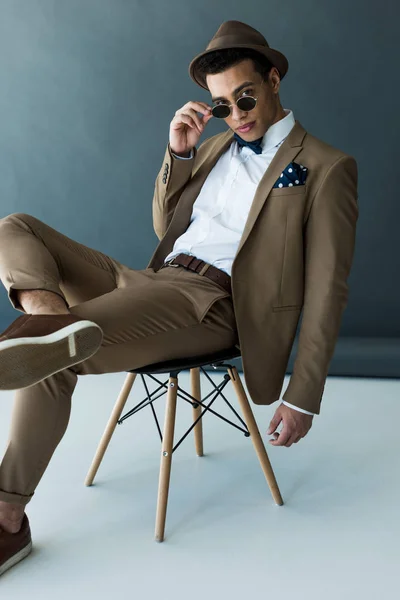 Elegante hombre de raza mixta en traje y gafas de sol sentado en la silla y mirando a la cámara en gris y blanco - foto de stock