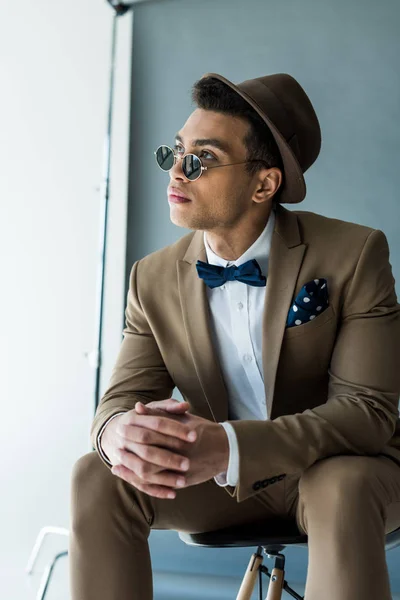 Stylish mixed race man in suit and sunglasses sitting on chair and looking away — Stock Photo