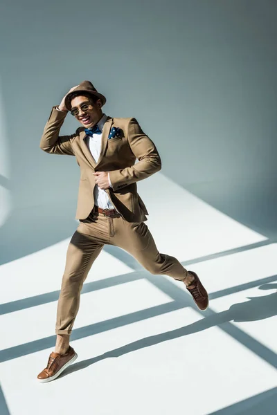 Homem de raça mista elegante em terno sorrindo e pulando em cinza com luz solar — Fotografia de Stock