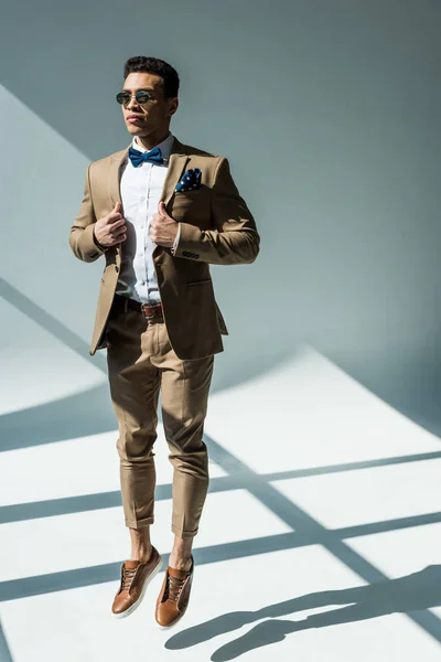 Elegante hombre de raza mixta en traje y gafas de sol saltando en gris con luz solar y espacio de copia - foto de stock