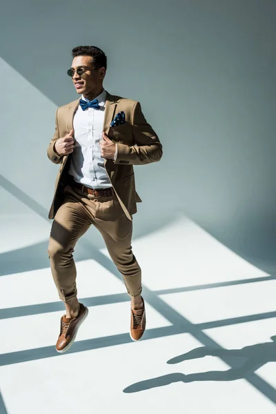 Stylischer Mixed Race Mann in Anzug und Sonnenbrille springt auf grau mit Sonnenlicht — Stockfoto