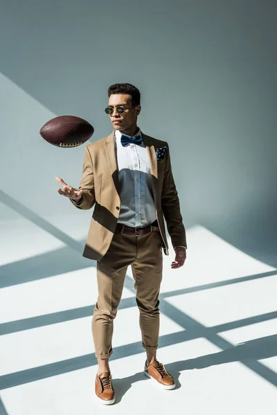 Elegante hombre de raza mixta en traje lanzando pelota de fútbol en gris con luz solar - foto de stock