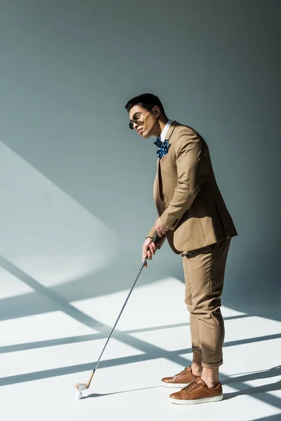 Stylish mixed race man in suit holding golf club on grey with sunlight — Stock Photo