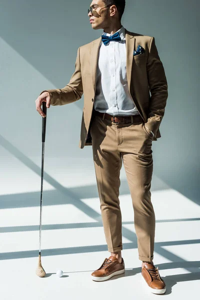 Elegante hombre de raza mixta en traje posando con club de golf en gris con la luz del sol - foto de stock