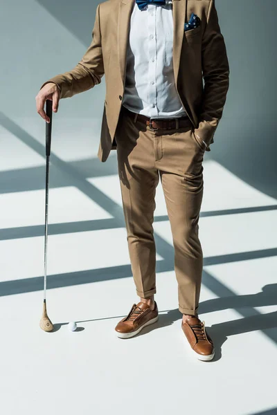 Vue recadrée de élégant homme de course mixte en costume posant avec club de golf sur gris avec lumière du soleil — Photo de stock