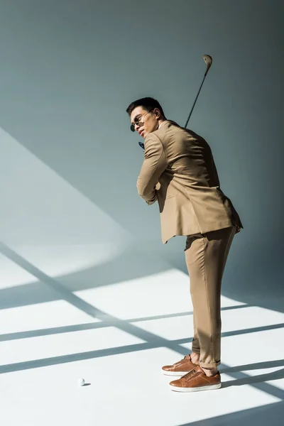 Elegante hombre de raza mixta en traje que sostiene el club de golf en gris con la luz del sol - foto de stock