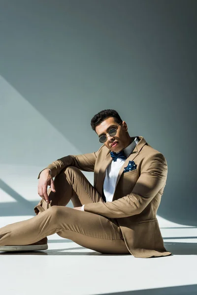 Stylischer Mixed Race Mann in Anzug und Sonnenbrille sitzt und posiert auf grau mit Sonnenlicht und Kopierraum — Stockfoto