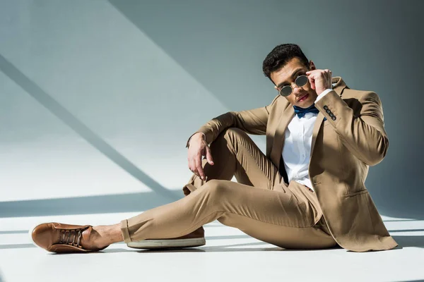 Stylish mixed race man in suit and sunglasses sitting and posing on grey with sunlight and copy space — Stock Photo