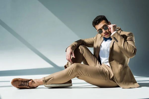 Élégant mixte homme en costume et lunettes de soleil assis et posant sur gris avec la lumière du soleil et l'espace de copie — Photo de stock