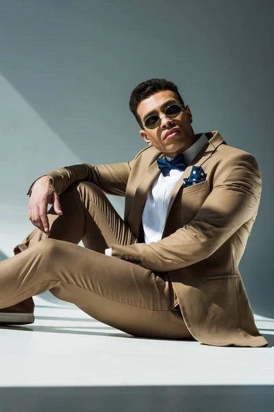 Elegante hombre de raza mixta en traje y gafas de sol sentado y posando en gris con luz solar - foto de stock