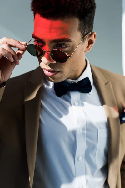 Élégant mixte homme en costume et lunettes de soleil posant sur gris avec lumière rouge — Photo de stock