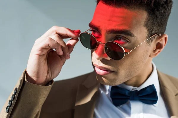 Élégant mixte homme en costume et lunettes de soleil posant sur gris avec lumière rouge — Photo de stock