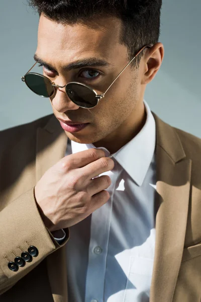 Stylish mixed race man in suit and sunglasses adjusting shirt and looking at camera on grey — Stock Photo