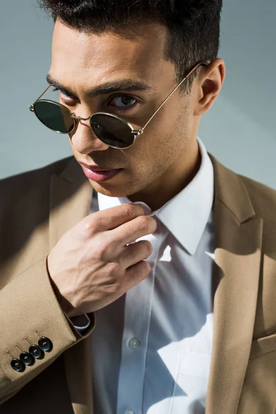 Elegante hombre de raza mixta en traje y gafas de sol ajustando la camisa y mirando a la cámara en gris - foto de stock