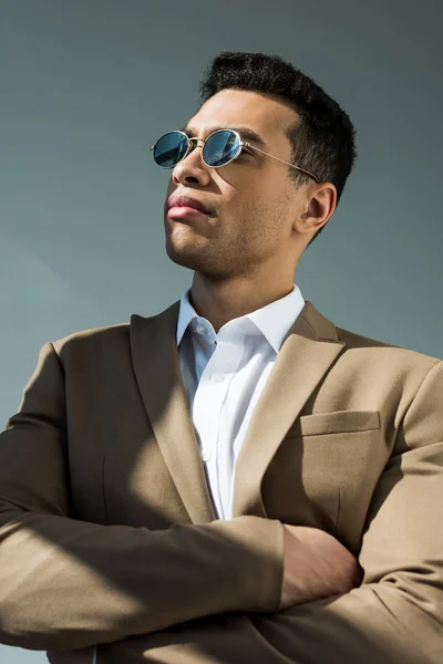 Stylischer Mixed Race Mann in Anzug und Sonnenbrille posiert mit verschränkten Armen auf grau mit Sonnenlicht — Stockfoto