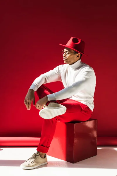 Stylish mixed race man in hat and glasses sitting and posing on red — Stock Photo