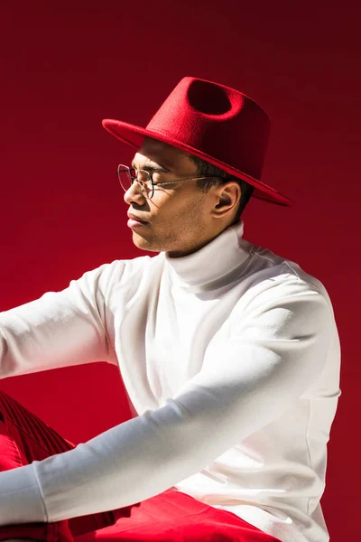 Selbstbewusster, stylischer Mischlingsmann mit Hut und Brille posiert isoliert auf Rot — Stockfoto